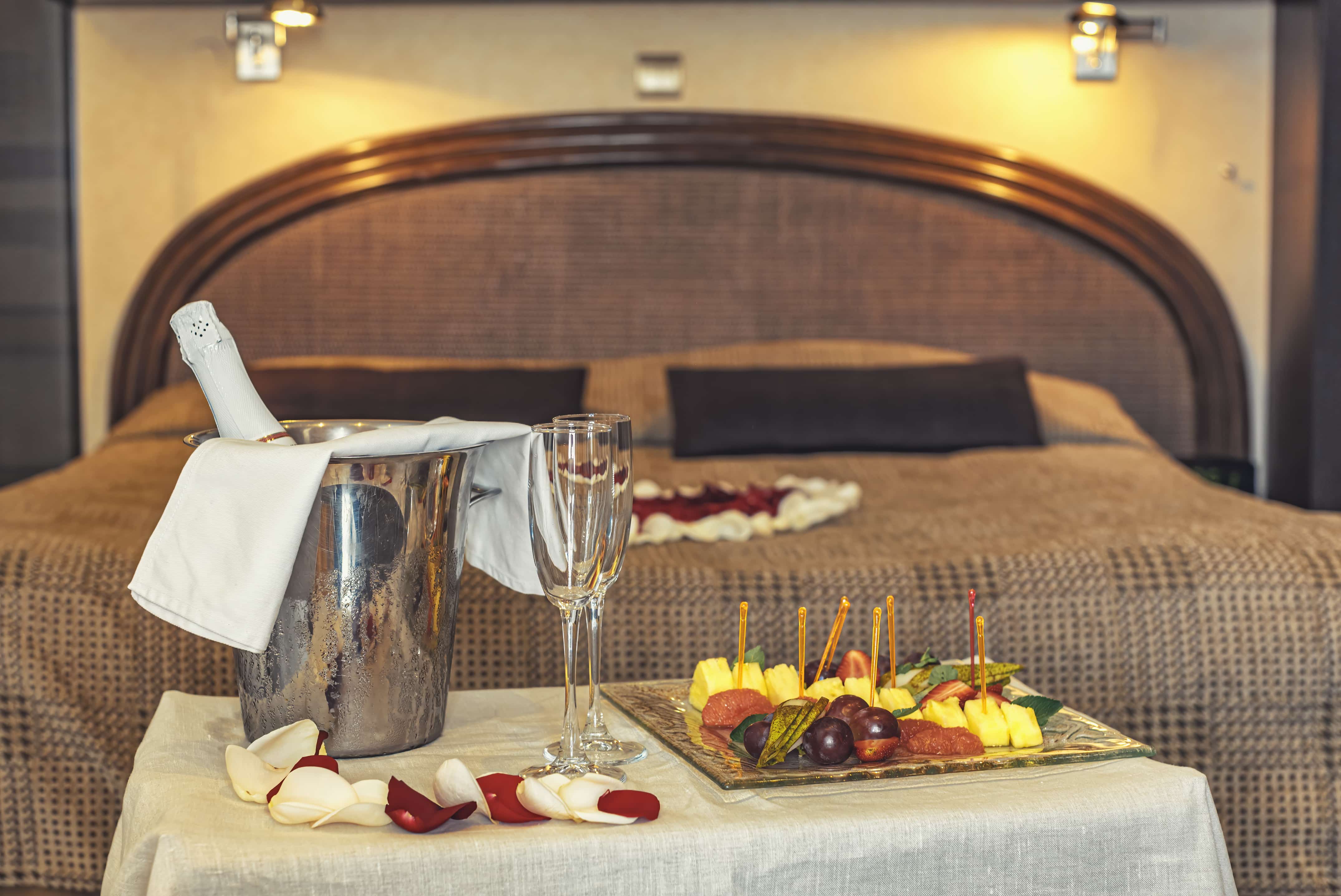 Elegant hotel room bed with welcome champagne in an ice bucket, 2 glasses, and a fruit platter.