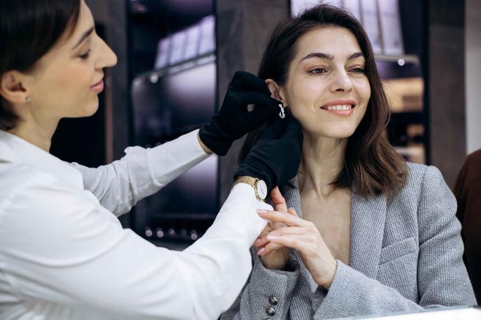 A female sales associate wearing a shirt and black gloves helps a customer try on some expensive earrings.