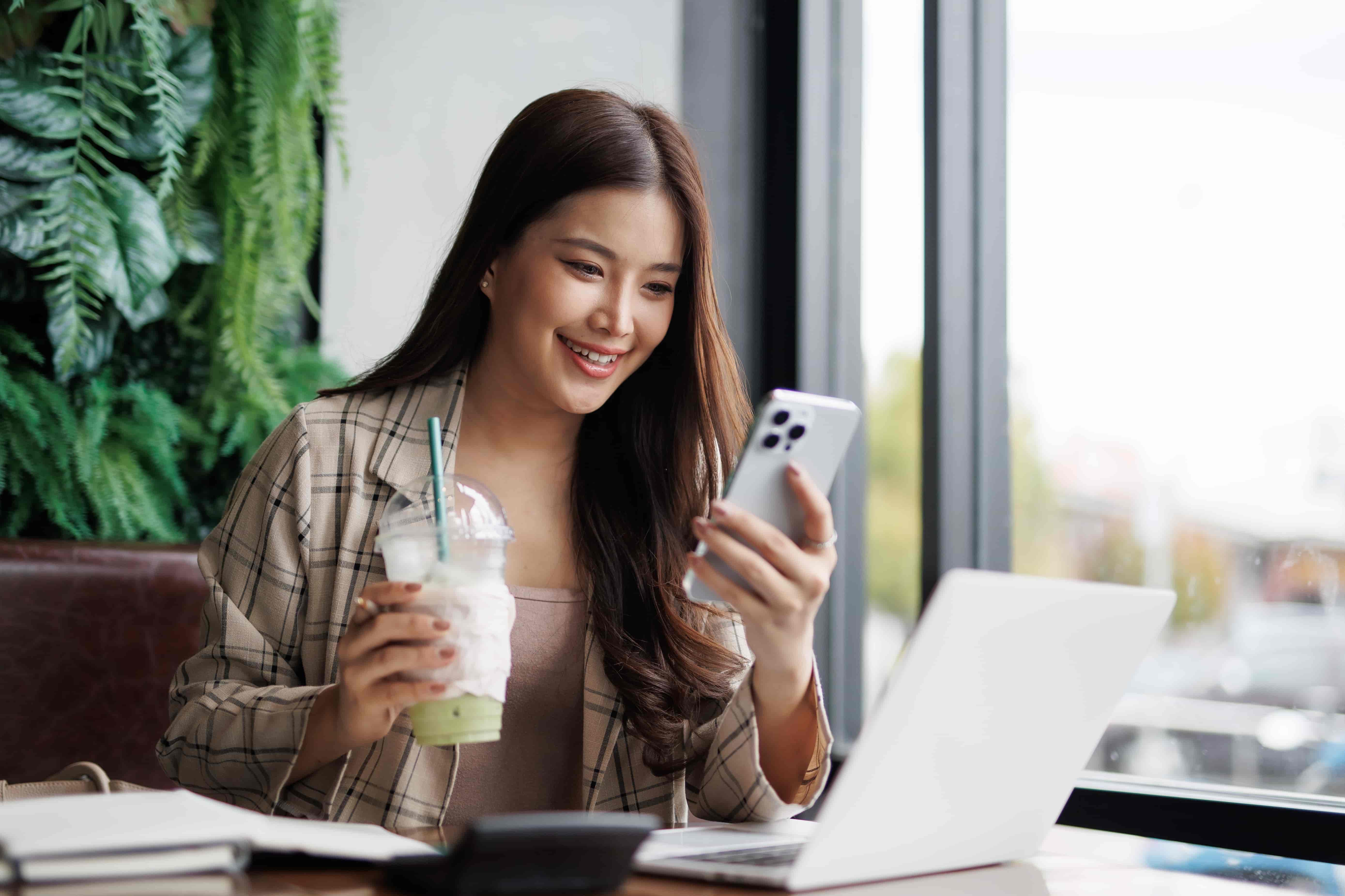 A woman in workwear, having a moment in a coffee shop with her smartphone, laptop and iced matcha drink