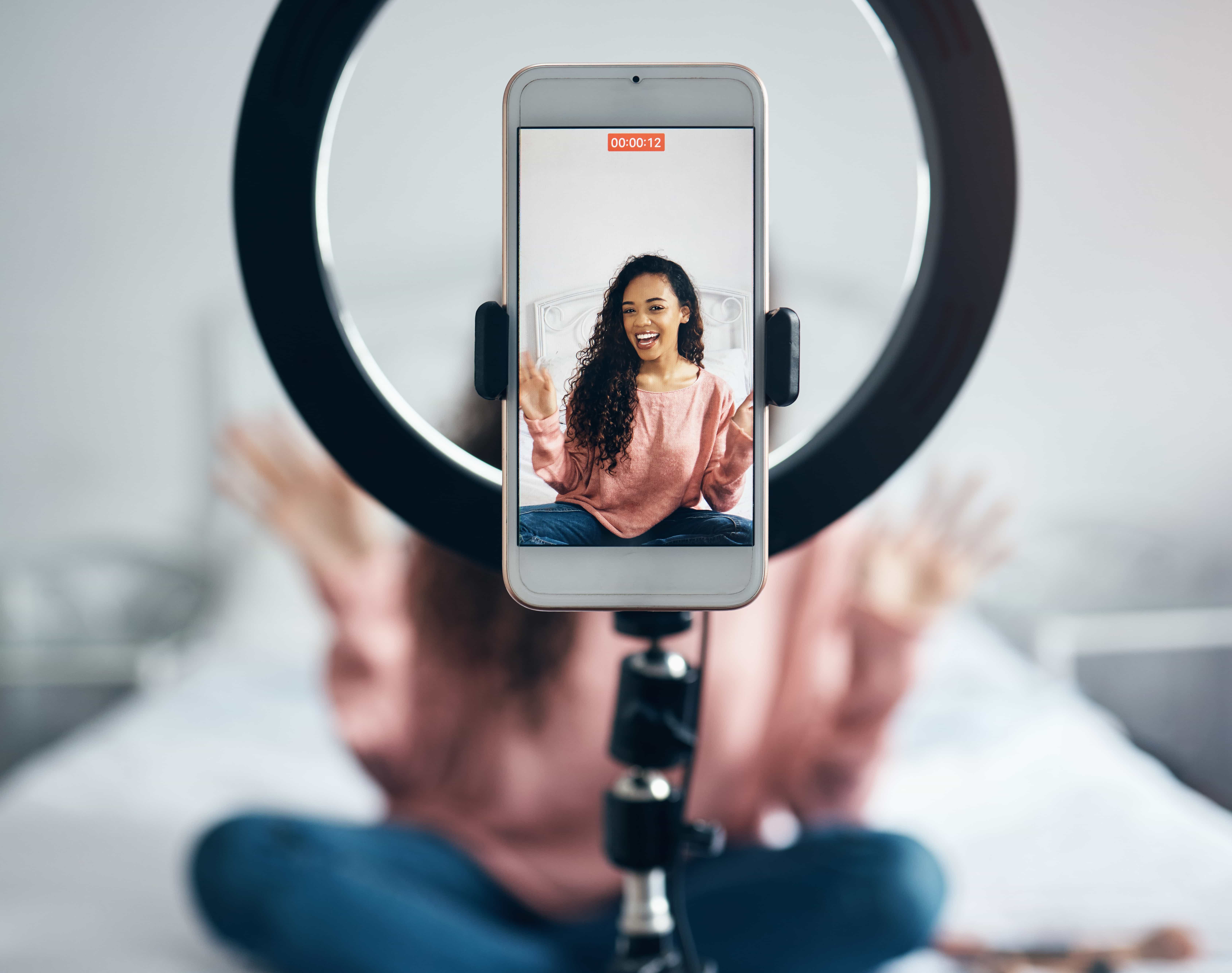 A social media influencer creating content on her bed with a ring light and smartphone set up on a tripod in front