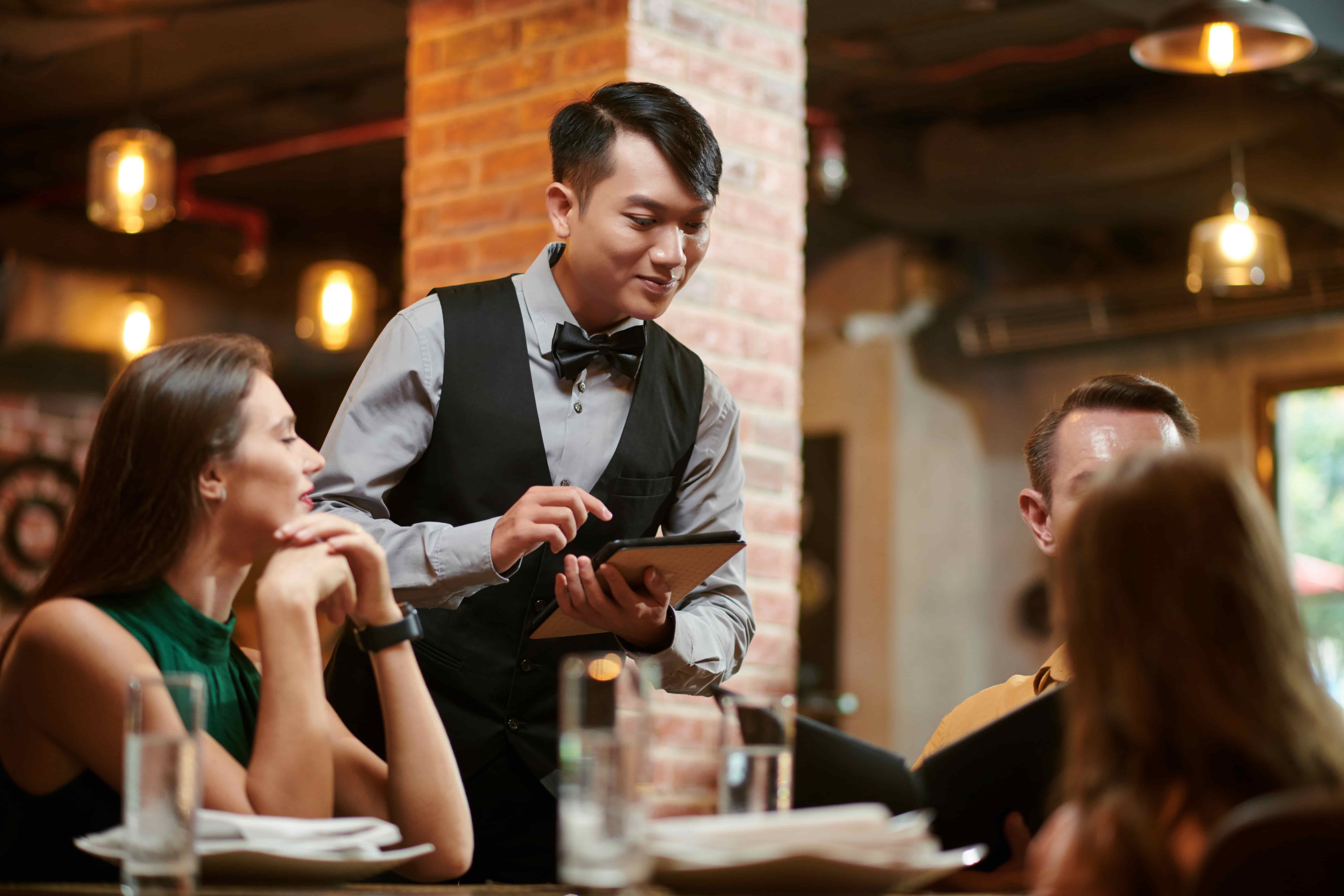 Waiter taking order on tablet for guests at a restaurant table with warm ambient lighting